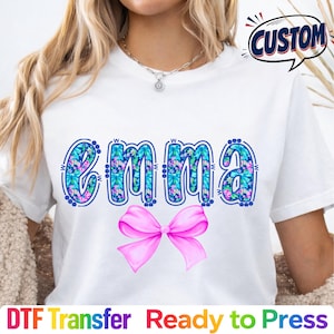 May include: White t-shirt featuring the name "EMMA" in a blue floral design, with a pink bow below. The word "CUSTOM" is in a speech bubble. The bottom of the shirt has the text "DTF Transfer Ready to Press".