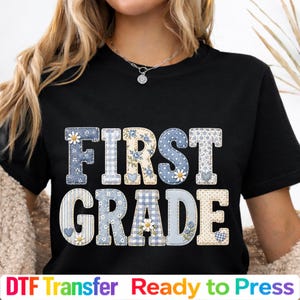 May include: A black t-shirt featuring the text FIRST GRADE in large block letters. The letters are filled with various blue and cream patterns, including gingham, floral prints, and small hearts, accented with white daisy flower graphics.