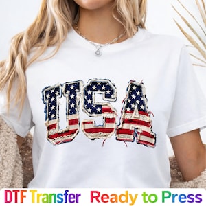 May include: White t-shirt with the word "USA" in a patriotic design. The letters are made with a denim-like fabric, red and white stripes, and blue fabric with white stars. The text "DTF Transfer Ready to Press" is at the bottom.