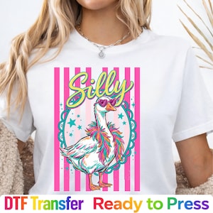 May include: White t-shirt featuring a cartoon goose wearing heart-shaped sunglasses and a colorful feather boa. The word "Silly" is written above the goose in a playful font. Pink and white striped background.