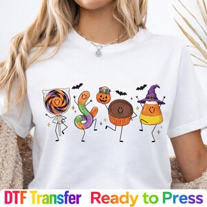 May include: A white t-shirt featuring a graphic print of four anthropomorphic Halloween treats: a swirled lollipop, a gummy worm, a chocolate peanut butter cup holding a jack-o'-lantern bucket, and a piece of candy corn wearing a witch hat.