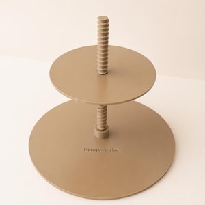 May include: A two-tiered, taupe-colored serving stand with a central, ridged support. The stand has two circular platforms, one smaller than the other. The text "Pingvicake" is printed on the lower tier.