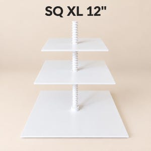 May include: A three-tiered white square display stand with a central threaded support rod. The text SQ XL 12" is printed at the top. The stand features three flat square platforms of varying sizes stacked vertically on the central column.