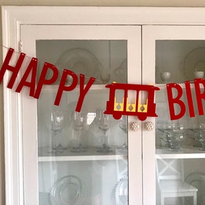 May include: A red felt banner with the words "HAPPY BIRTHDAY" strung across a thin silver cord. A red train car with yellow accents is between the words. The banner is hanging in front of a white cabinet with glass doors.