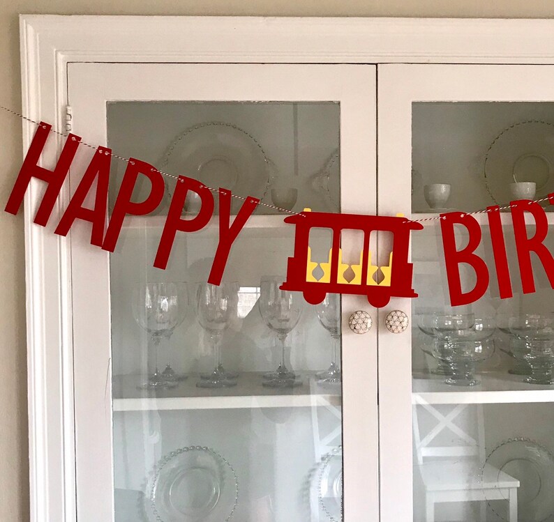 May include: A red felt banner with the words "HAPPY BIRTHDAY" strung across a thin silver cord. A red train car with yellow accents is between the words. The banner is hanging in front of a white cabinet with glass doors.