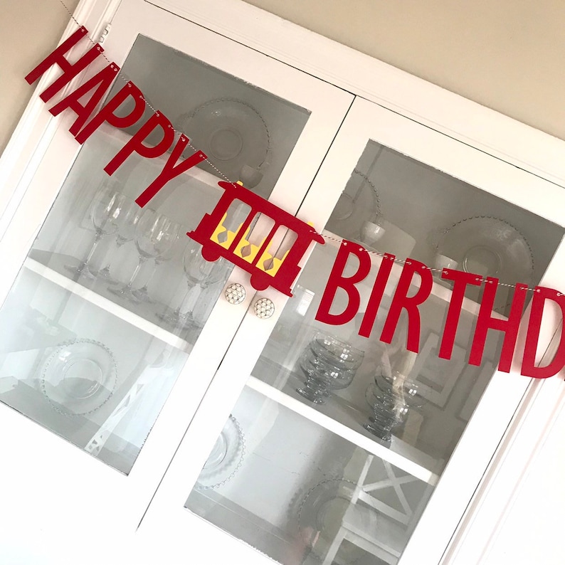 May include: Red "HAPPY BIRTHDAY" banner with a train illustration. The banner is strung across a white cabinet with glass doors, showcasing glassware inside. The train is red with yellow accents.
