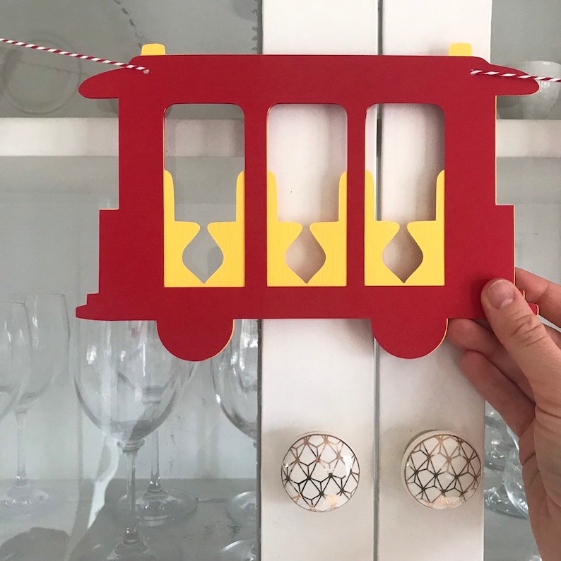 May include: A red and yellow paper train car decoration with three window cutouts. The train car has a decorative shape and is suspended by a red and white string. The train car is held by a hand.