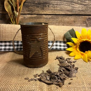 May include: A cylindrical, rusted metal container featuring a star-shaped cutout with a burlap backing and a thin twine handle. The container is positioned near a bright yellow sunflower and a small pile of dark wood chips on a textured surface.