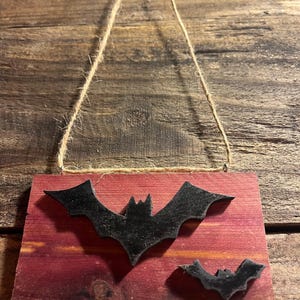 Puede incluir: Una placa rectangular de madera teñida en un tono marrón rojizo profundo, con dos siluetas de murciélagos negros adheridas al frente. La placa está suspendida por un lazo de cuerda de yute de color natural sujeto al borde superior para colgar.