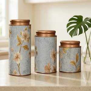 May include: A set of three decorative canisters with copper-coloured lids. The canisters have a light grey background with a floral design in beige and gold. The canisters are cylindrical and vary in size.