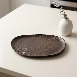 May include: A dark brown, oval-shaped ceramic serving platter with a textured surface and a floral pattern along the edge. A small white vase with a sprig of greenery is in the background.