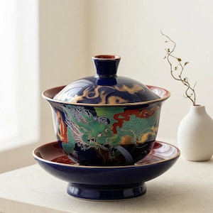 May include: A navy blue ceramic tea set with a dragon design. The set includes a lidded bowl, a saucer, and a base. The dragon is green, red, and gold. The lid and the rim of the bowl are gold.