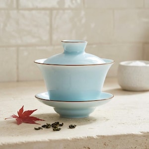 May include: A light blue porcelain gaiwan tea set with a matching lid and saucer. The tea set has a brown trim and sits on a light-colored surface. A red maple leaf and loose tea leaves are next to the tea set.
