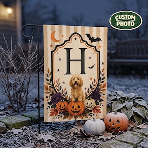 May include: A rectangular garden flag with a beige and white striped background featuring a central black letter H, a golden doodle dog, orange pumpkins, purple roses, autumn leaves, a crescent moon, and bats. The flag is on a black metal stand.