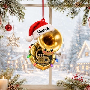 Peut inclure: Un ornement en forme de tuba doré arborant un visage souriant aux grands yeux, coiffé d'un bonnet de Père Noël rouge et blanc avec le mot Sweetie inscrit sur le rebord. L'instrument est entouré d'une guirlande lumineuse et suspendu par un ruban rouge.