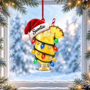 Op de afbeelding: Een hangend ornament in de vorm van een glas gele ananassap, bekroond met een rode kerstmuts met de tekst Sweetie. Het glas is omwikkeld met een snoer van kleurrijke kerstverlichting en is gegarneerd met een schijfje ananas.