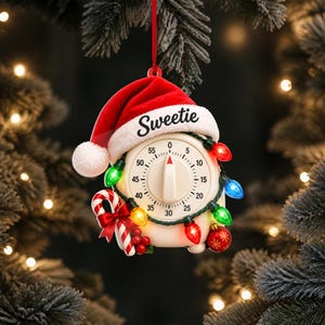 Peut inclure: Une décoration de Noël en forme de minuteur de cuisine portant un bonnet de Père Noël rouge avec le mot Sweetie inscrit sur le rebord blanc. Il est orné d'une guirlande lumineuse colorée, d'un sucre d'orge avec un nœud rouge et d'une boule.