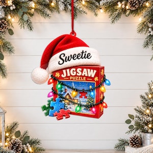 May include: A hanging Christmas ornament shaped like a red jigsaw puzzle box with the word Sweetie on a Santa hat. The box features a winter scene, loose blue and red puzzle pieces, and a string of colorful glowing Christmas lights wrapped around.