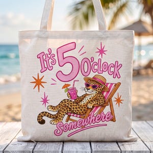 Puede incluir: Un bolso de tela color crema con el gráfico de un leopardo con sombrero de paja y gafas de sol rosas, descansando en una silla de playa mientras sostiene un cóctel rosa. El texto dice It's 5 O'clock Somewhere en letras de color rosa.