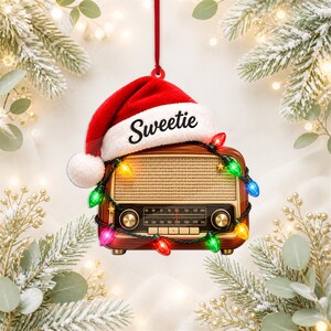 Könnte beinhalten: Ein weihnachtlicher Anhänger in Form eines Vintage-Radios mit einer roten Weihnachtsmannmütze, auf deren weißem Rand das Wort Sweetie steht. Das Radio ist mit einer Lichterkette aus bunten, leuchtenden Weihnachtslichtern umwickelt.