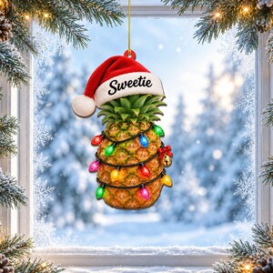 May include: A hanging pineapple ornament featuring a red and white Santa hat with the word Sweetie printed on the brim. The pineapple is wrapped in a string of colourful Christmas lights in red, green, blue, yellow, and pink, with a small gold bell.