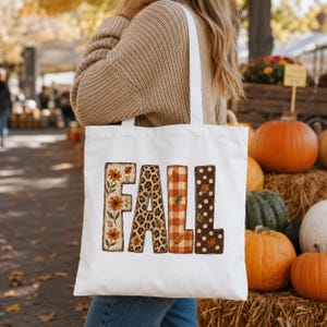May include: A white canvas tote bag featuring the word FALL printed in large block letters. Each letter has a distinct pattern: the F has orange flowers, the A has a leopard print, the L has orange plaid, and the L has brown polka dots.