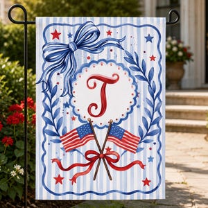 May include: A patriotic garden flag featuring a blue bow, red stars, and two American flags. The flag has a white center with a red letter "T" and is framed by blue leaves and a scalloped border. The background has blue and white stripes.