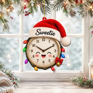 Op de afbeelding: Een rond klokornament met een rode kerstmuts met de tekst Sweetie op de witte rand. De wijzerplaat heeft een lachend gezichtje, zwarte wijzers en is versierd met een snoer van gekleurde kerstverlichting en een kleine rode strik.
