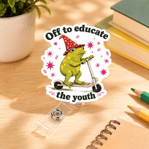 May include: A badge reel featuring a die-cut sticker of a green frog wearing a red wizard hat with yellow stars, riding a silver scooter. The text "Off to educate the youth" is printed in dark green letters above and below the frog illustration.