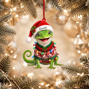 Op de afbeelding: Een hangend ornament met een groene hagedis die een rood-witte kerstmuts draagt met het woord Sweetie op de rand gedrukt. De hagedis is gekleed in een rood-groene trui met patroon, versierd met een snoer van kleurrijke kerstverlichting.