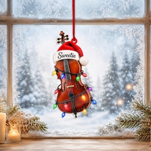 Könnte beinhalten: Ein brauner Geigenanhänger aus Holz, der an einem roten Band hängt, mit einer roten Weihnachtsmannmütze, auf deren weißem Rand das Wort Sweetie gedruckt ist, umwickelt mit einer Lichterkette mit roten, blauen, grünen und gelben Lichtern.