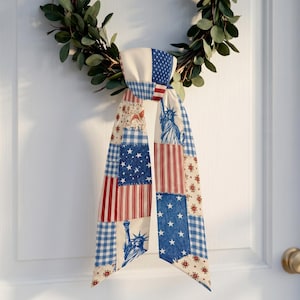 Peut inclure: Un nœud de porte patriotique avec un motif patchwork représentant le drapeau américain, des étoiles et la Statue de la Liberté. Le nœud est attaché à une couronne verte, créant un accent festif et décoratif pour une porte d'entrée.