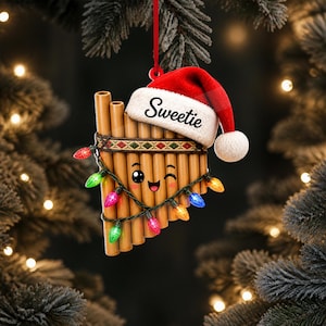 Puede incluir: Un adorno de flauta de pan con una cara sonriente y guiñando un ojo con mejillas rosadas, que lleva un gorro de Papá Noel rojo y blanco con la palabra Sweetie impresa en el borde. El instrumento está envuelto en luces navideñas de colores y cuelga de una cinta roja.
