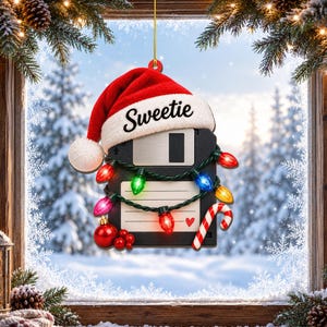 May include: A black floppy disk ornament wearing a red and white Santa hat with the word Sweetie printed on the brim. The disk is wrapped in a string of colourful Christmas lights, a red ornament ball, and a candy cane with a small red heart icon.