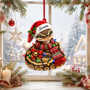 May include: A hanging Christmas ornament featuring a ball python wearing a red Santa hat with the word Sweetie printed on the brim, a red and green patterned scarf, and a gold bell with a red bow, wrapped in a string of colorful holiday lights.