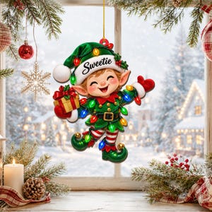 May include: A hanging Christmas ornament shaped like a smiling elf wearing a green hat with the word Sweetie printed on the white brim. The elf wears a green outfit with a gold belt, red and white striped leggings, and holds a gold gift box.