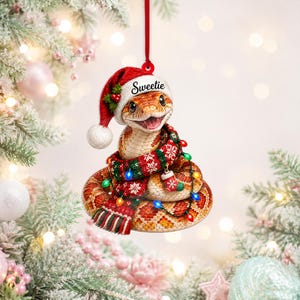 Könnte beinhalten: Ein hängender Weihnachtsschmuck in Form einer zusammengerollten Schlange, die eine rote Weihnachtsmütze mit dem Wort Sweetie auf dem Rand trägt. Die Schlange ist in einen rot-grünen Schal mit Muster gewickelt und mit einer kleinen Socke verziert.