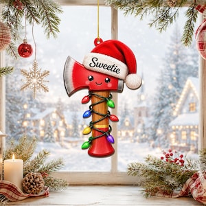 Könnte beinhalten: Ein hängendes Ornament in Form einer roten Axt mit einem lächelnden Gesicht, das eine rot-weiße Weihnachtsmütze mit dem Wort Sweetie auf dem Rand trägt. Der Holzgriff ist mit einer Lichterkette aus bunten Weihnachtsleuchten umwickelt.