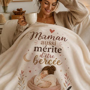 Può includere: Una coperta color crema con il testo francese "Maman aussi mérite d'être bercée" e una grafica di una donna che abbraccia un bambino. La coperta ha un design a cuore e stella. Una donna tiene in mano una tazza.