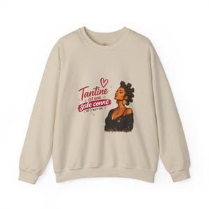 Peut inclure: Sweat-shirt beige avec un imprimé d'une femme portant des boucles d'oreilles dorées et un collier. Le texte "Tantine est une sale conne et c'est ok" est imprimé en rouge. Le sweat-shirt a un col rond et des manches longues.