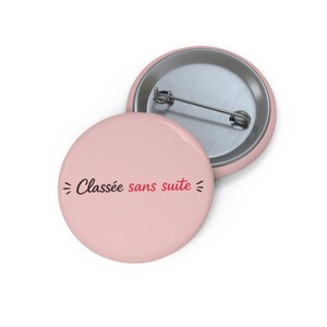 Peut inclure: Deux badges ronds rose clair. L'un des badges, au premier plan, affiche le texte "Classée sans suite" en script noir et rouge. L'autre badge est partiellement visible à l'arrière-plan.