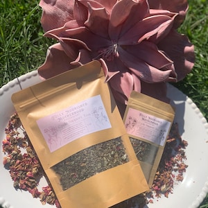 May include: Two brown paper pouches of Cycle Support Tea sit on a white plate with a decorative edge, surrounded by dried rose petals. A large, dusty rose-colored flower sculpture is in the background. The pouches have the text "Fully Nourished Motherhood" and "Cycle Support Tea".