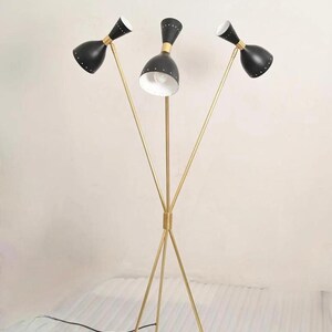 May include: A mid-century modern floor lamp with three black cone-shaped shades, each with a white interior. The shades are attached to gold-colored metal stems that converge at the base. The lamp has a tripod base.