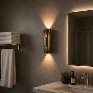 Könnte beinhalten: Eine vertikale Wandleuchte mit metallischem Bronze-Finish und einem geschwungenen, überlappenden Design. Die Leuchte strahlt warmes, fokussiertes Licht aus den oberen und unteren Öffnungen aus und wirft einen sanften Schein auf die geflieste Wand.