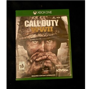 May include: A Call of Duty WWII video game case for Xbox One. The cover features a soldier in a helmet with hands covering the face, holding dog tags. Text includes Call of Duty WWII, 4K Ultra HD, HDR, Sledgehammer Games, Activision, and ESRB M.