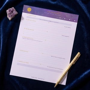 Pode incluir: Um Daily Soul Journal em uma superf&iacute;cie de veludo azul escuro, com uma caneta dourada e um cristal roxo. O di&aacute;rio tem um cabe&ccedil;alho roxo e branco com o t&iacute;tulo "Daily Soul Journal". O di&aacute;rio tem prompts para reflex&atilde;o.
