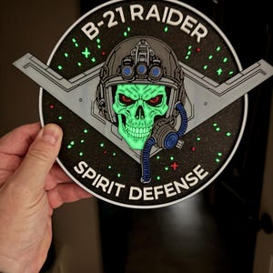 May include: A circular patch featuring a glowing green skull wearing a pilot helmet with a blue oxygen mask. The background is black with green star-like accents and a gray aircraft silhouette. Text reads B-21 RAIDER and SPIRIT DEFENSE.