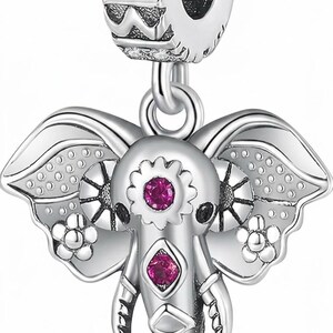 Puede incluir: Un dije de plata con forma de elefante y orejas grandes, con dos pequeñas piedras rosas incrustadas en la frente y la trompa. El pasador cilíndrico superior tiene piedras transparentes y texto grabado, conectando con el colgante inferior.
