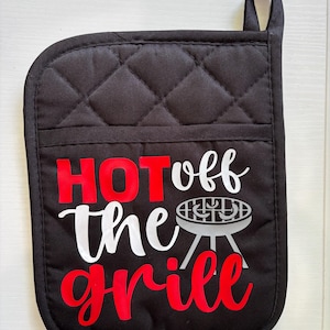May include: A black quilted oven mitt with a pocket, featuring the red and white text "HOT off the grill" and a graphic of a charcoal grill with two sausages on the grate. The mitt includes a small fabric loop at the top corner for hanging.
