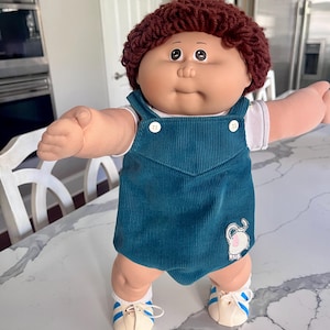 Puede incluir: Una muñeca vintage con cara redonda, cabello de hilo marrón y ojos grandes. La muñeca viste un peto de pana verde azulado con botones blancos y un pequeño parche de elefante. Tiene calcetines blancos y zapatos color crema con rayas azules.
